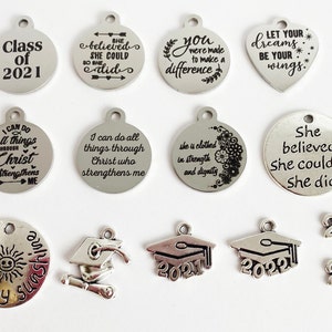 May include: A collection of silver charm pendants with various graduation-themed designs and inspirational quotes. The charms include a graduation cap, a diploma, the year 2021, and quotes such as "She believed she could so she did" and "You were made to make a difference."