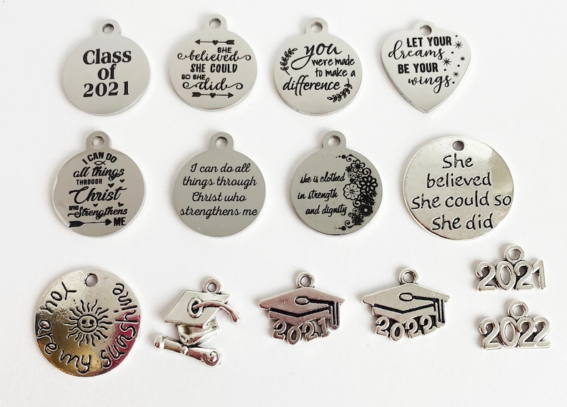 May include: A collection of silver charm pendants with various graduation-themed designs and inspirational quotes. The charms include a graduation cap, a diploma, the year 2021, and quotes such as "She believed she could so she did" and "You were made to make a difference."