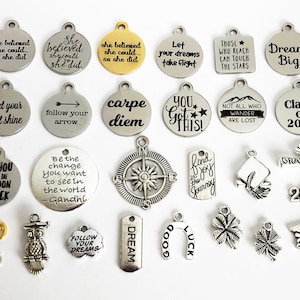 May include: A collection of silver and gold charm pendants with inspirational quotes and graduation themes. The charms feature phrases like "Let your light shine", "Dream Big", "Follow your arrow", "Carpe Diem", "You Got This!" and "Class of 2019".