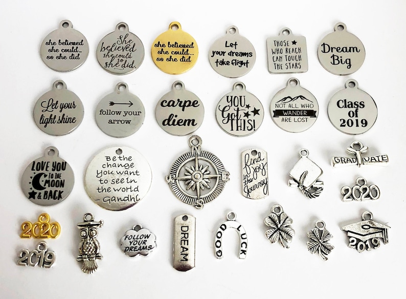 May include: A collection of silver and gold charm pendants with inspirational quotes and graduation themes. The charms feature phrases like "Let your light shine", "Dream Big", "Follow your arrow", "Carpe Diem", "You Got This!" and "Class of 2019".