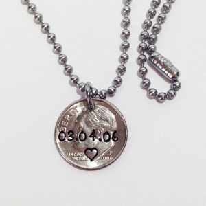 May include: A silver-toned necklace with a ball chain and a coin pendant. The coin pendant features the date "03.04.06" and a heart symbol. The necklace is displayed against a white background.