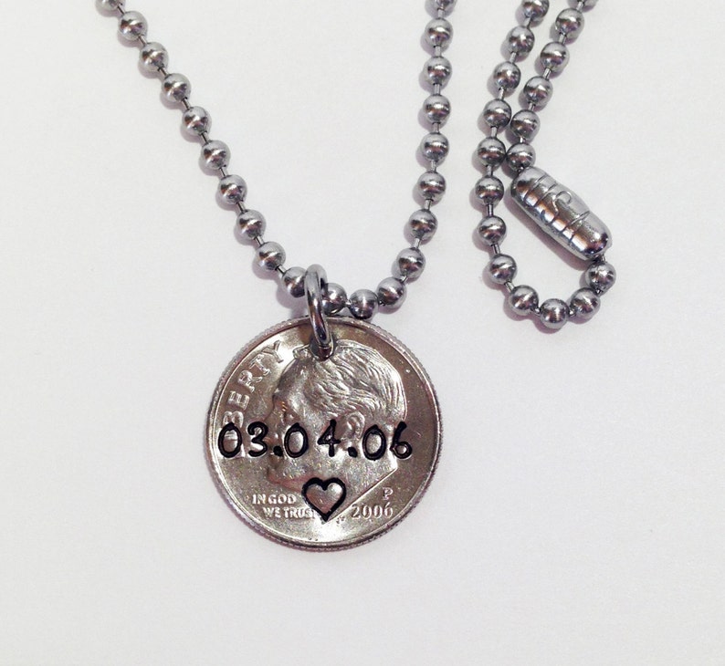 May include: A silver-toned necklace with a ball chain and a coin pendant. The coin pendant features the date "03.04.06" and a heart symbol. The necklace is displayed against a white background.