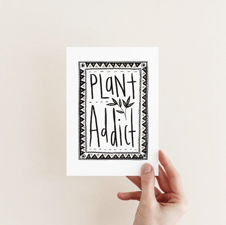 May include: A white card with the words "PLANT ADDICT" in black, handwritten-style lettering. The text is inside a decorative black and white border. A small sprig of leaves is between the words. The card is held by a hand.