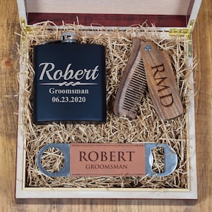 May include: A wooden gift box with a black flask, a wooden comb, and a bottle opener. The flask is engraved with the name "Robert" and the date "06.23.2020". The comb is engraved with the initials "RMD". The bottle opener has a leather patch with the name "Robert" and the word "Groomsman".