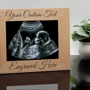 May include: A wooden picture frame with a light brown finish. The frame has a white mat and is engraved with the text "Your Custom Text" at the top and "Engraved Here" at the bottom. The frame is holding a black and white ultrasound image of a baby.
