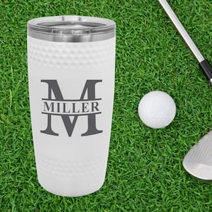 May include: White stainless steel tumbler with a golf ball pattern. The tumbler has a grey monogram with the letter "M" and the name "Miller" printed on it.