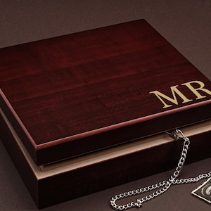May include: A dark wood box with a hinged lid. The lid is engraved with the initials "MR" in gold lettering. The box is open and a silver chain is hanging out of it.