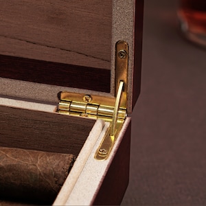 May include: Close-up of a wooden box with a brass hinge. The box is open and has a brown interior. The box is made of dark wood with a light wood inlay.