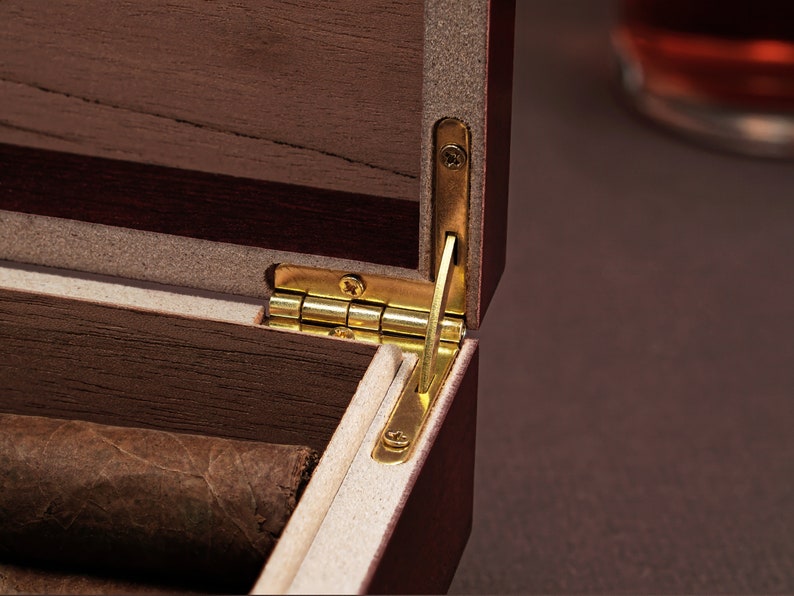 May include: Close-up of a wooden box with a brass hinge. The box is open and has a brown interior. The box is made of dark wood with a light wood inlay.