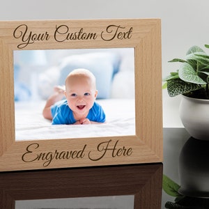 May include: A light wood picture frame with a baby photo inside. The frame has the text "Your Custom Text" at the top and "Engraved Here" at the bottom.