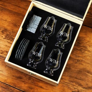 May include: A wooden box with a black interior contains four clear glass whiskey glasses with the initials "W/M" etched on each. The box also includes four black coasters and a bag of grey ice cubes.