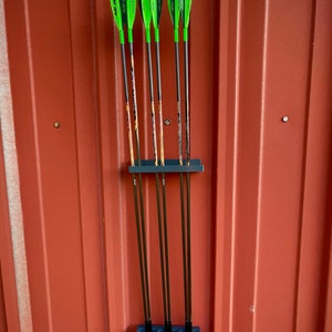May include: A gray arrow holder with six arrows. The arrows have green fletching and black shafts. The holder is mounted on a red wall.