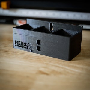 May include: A black, 3D-printed organizer with three compartments and two circular holes. The organizer has the text "MORR INNOVATIONS 1.0" on the side. It is sitting on a wooden surface.