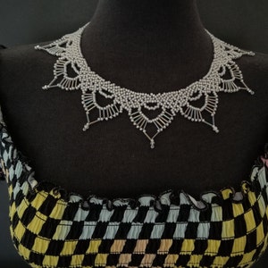 May include: A silver beaded necklace with a delicate, lacy design. The necklace is displayed on a black and white checkered fabric background.