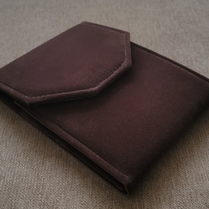 May include: A brown velvet jewelry pouch with a flap closure. The pouch is folded and lying on a textured surface.