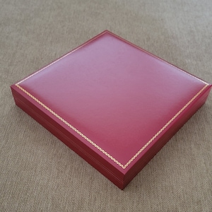 May include: A red square box with a gold trim around the edges. The box is made of leather and has a smooth surface.