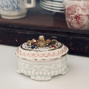 May include: A small, white porcelain trinket box with a lid. The lid is decorated with gold trim, red and blue floral accents, and a gold-colored handle. The box has a detailed, textured design around the base. Other antique porcelain pieces are in the background.