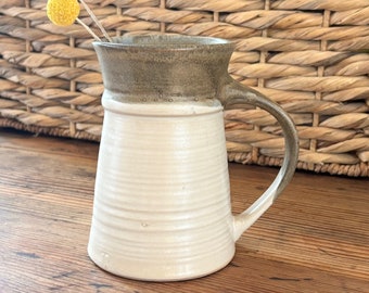 May include: A white ceramic pitcher with a brown glaze on the top rim and handle. The pitcher has a slightly flared lip and a rounded base. It is sitting on a wooden surface.