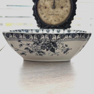 May include: A white and blue floral patterned ceramic bowl. The bowl has a rectangular shape with curved sides and a decorative border. The floral design features dark blue flowers and leaves. An antique clock is in the background.