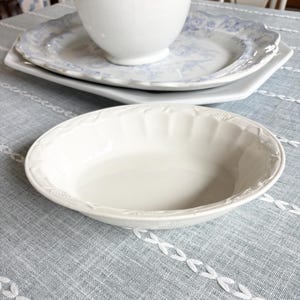 May include: A white ceramic bowl with a decorative rim sits on a light blue tablecloth. In the background, a white pitcher rests on a stack of plates with a blue floral design. The scene suggests a dining or kitchen setting.