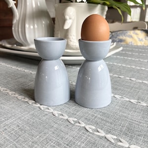 May include: Two light blue ceramic egg cups. One cup holds a brown egg. The egg cups have a rounded base and a small, shallow cup on top. The background includes a gray tablecloth and white decorative vases.