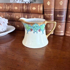 May include: A small, antique cream pitcher with a gold handle and rim. The pitcher has a cream-colored base and is decorated with green, blue, and red floral designs. The pitcher is sitting on a dark wooden surface, with books in the background.