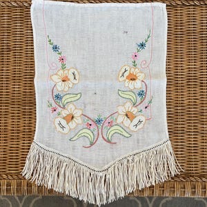 May include: Cream-colored vintage table runner with embroidered floral design. Features large white flowers with orange centers, green leaves, and pink, blue, and yellow accents. The bottom edge has a fringe.
