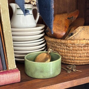 May include: A vintage still life arrangement featuring a green ceramic bowl with a gold pocket watch, a stack of white plates, a white pitcher with green stripes, and a woven basket. A blue ribbon and old books add to the antique aesthetic.