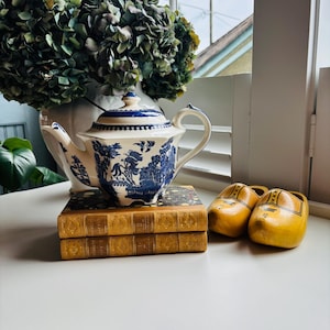 May include: A blue and white porcelain teapot with a detailed pattern sits atop two antique books. A pair of yellow wooden clogs rests nearby. A large bouquet of dried hydrangeas is in the background.