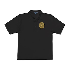 Peut inclure: Un polo noir à manches courtes avec un col, une patte de boutonnage à trois boutons et un emblème circulaire doré sur la poitrine gauche. L'emblème présente un motif géométrique central entouré de lauriers et le texte PROMPT ENGINEER.
