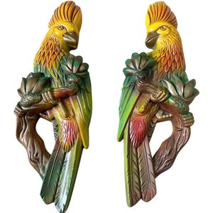 May include: Pair of hand-painted parrot wall sculptures. Each parrot is perched on a branch, with yellow, green, and red plumage. The parrots are holding a flower in their claws. The sculptures are made of a durable material.