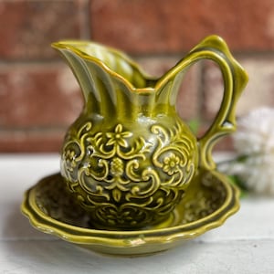 May include: A green ceramic pitcher and saucer set. The pitcher has a handle and a spout, and both pieces feature a floral relief pattern. The set is displayed on a white surface with a blurred brick background.