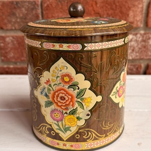 May include: A vintage, round tin with a lid, featuring a floral design in shades of orange, yellow, and pink against a brown background. The tin has a wooden appearance and a small, round knob on top. The background is a brick wall.