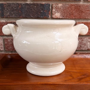May include: A cream-colored ceramic bowl with a rounded body, a pedestal base, and two scrolled handles on the sides. The bowl features a smooth, glossy finish and is positioned in front of a fabric item with pink floral and butterfly patterns.