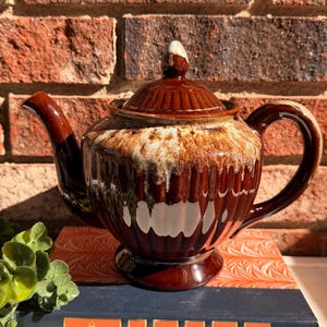 May include: A vintage brown ceramic teapot with a dripped glaze design, resting on a book. The teapot has a lid and handle. The book has the title "Reader's Digest Condensed Books" on the spine.