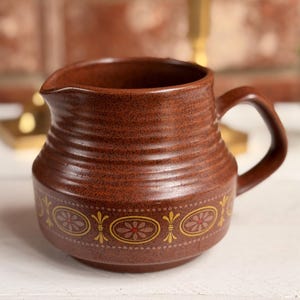 May include: A brown ceramic pitcher with a handle and a spout. The pitcher has horizontal ridges and a decorative band of yellow and red floral designs around the base. The background includes a brick wall and gold candle holders.