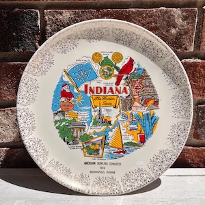 May include: A white ceramic plate with a gold rim and a colorful illustration of the state of Indiana. The plate features landmarks, symbols, and text that reads "The Hoosier State" and "American Bowling Congress 1954 Indianapolis, Indiana".