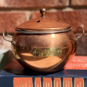 May include: A round copper potpourri container with two side handles and a removable lid featuring small ventilation holes. A brass plaque on the front is engraved with the word POTPOURRI. The container rests on a blue and orange book.