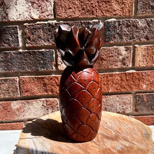 May include: A hand-carved wooden pineapple sculpture with a dark brown finish. The pineapple is detailed with a textured surface and a leafy crown.