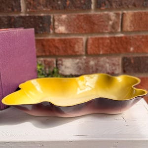 May include: A vintage, leaf-shaped serving dish with a gradient of colors. The dish transitions from a bright yellow interior to a deep burgundy exterior. The dish is placed on a white surface with a brick background.