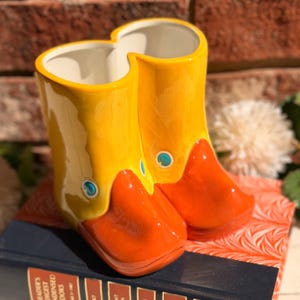 May include: A ceramic duck boot planter in yellow and orange, with blue accents, sits atop a vintage book. The planter has a whimsical design, perfect for adding a touch of fun to any space.
