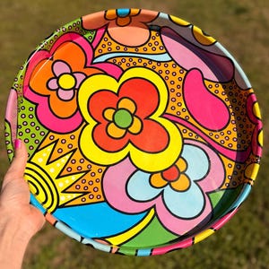 May include: A round, colorful tray with a vibrant floral design. The tray features large, stylized flowers in shades of orange, yellow, pink, and blue, set against a background of smaller dots. The tray's rim is a bright pink.