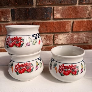 May include: White ceramic soup bowls with the Campbell's logo and illustrations of tomatoes, carrots, and other vegetables. The bowls have a blue and white checkered border and are stacked on a white surface.