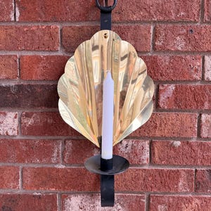 May include: A wall-mounted candle holder with a brass-colored, fan-shaped backplate. A white candle sits in a black holder. The holder is attached to a black metal bracket, hanging against a red brick wall.
