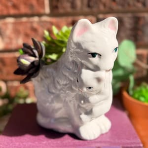 May include: A white ceramic cat planter with a kitten nestled against its chest. The cat has pink-lined ears and blue eyes. The planter holds a succulent plant with green and purple leaves. The planter is sitting on a purple book.