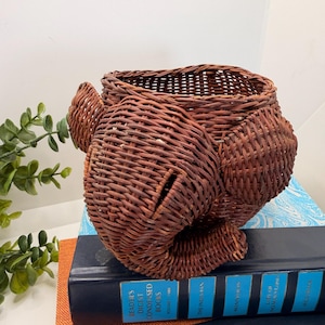 May include: A woven brown basket with a unique elephant design, featuring a trunk and ears. The basket is made of tightly woven wicker and sits atop a stack of books. The basket's color is a warm, reddish-brown.