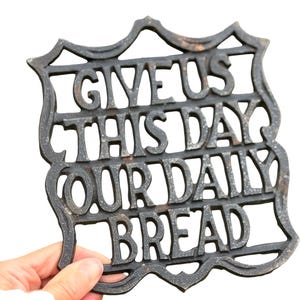May include: A dark gray cast iron trivet with a shield-like shape and a textured surface. The metal piece features the raised, embossed text "GIVE US THIS DAY OUR DAILY BREAD" arranged in four horizontal lines within the decorative frame.