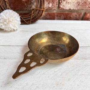 May include: A small, brass, vintage serving dish with a handle. The dish has a round bowl and a long, thin handle with decorative cutouts.