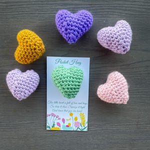 May include: Five crocheted heart-shaped stuffed toys in various colors, including purple, pink, green, and yellow. The hearts are arranged around a card that reads "Pocket Hug" and includes a message about love and hugs.