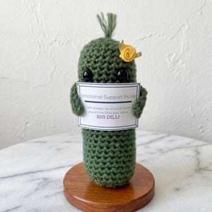 May include: A handmade, green crocheted pickle with black eyes and a small yellow flower. The pickle holds a sign that reads "Emotional Support Pickle. I will always be around to let you know that you are a BIG DILL!" It sits on a wooden base.